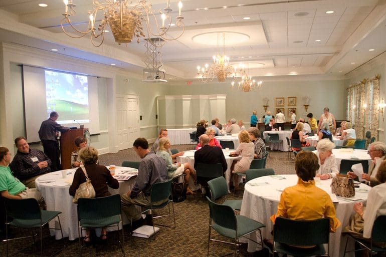 A group of people sitting at tables in front of a projector screen.