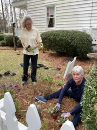 Two women planting flowers by white picket fence