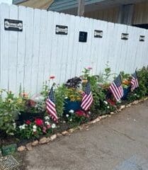 A white fence with flowers and flags in the middle of it.