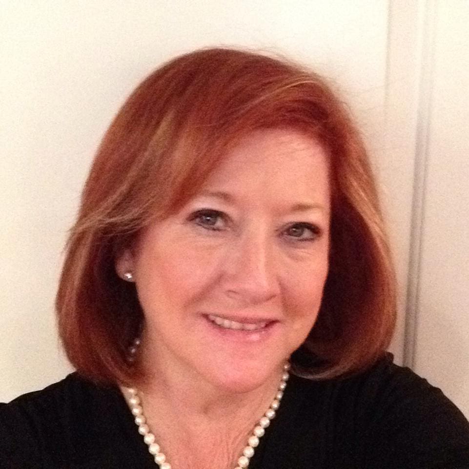 A smiling woman with red hair wearing a black top and pearl necklace.