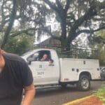 A man smiling near a white utility truck under large trees.